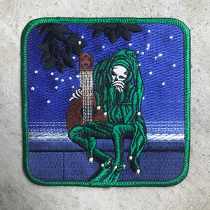 May include: Embroidered patch featuring a green jester figure holding a guitar, set against a blue starry night background. The jester is wearing a green hat and has a white face.