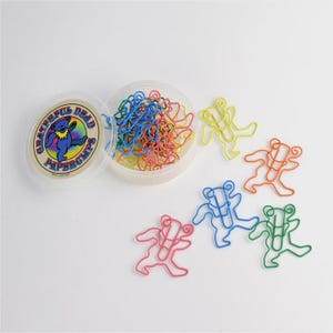 May include: A clear plastic container with a lid featuring a Grateful Dead logo, next to a pile of colorful bear-shaped paperclips. Several individual paperclips in yellow, orange, blue, green, and pink are scattered around.