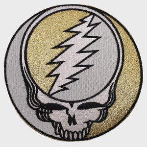May include: Embroidered patch featuring a stylized skull with a lightning bolt on a gold and silver background. The patch has a black border and is circular in shape. Ideal for jackets, bags, or other fabric items.
