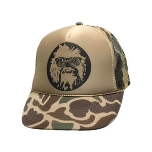 May include: A tan and camouflage trucker hat with a black and white graphic of a furry creature wearing sunglasses and the text "WIDE SPREAD PANIC".