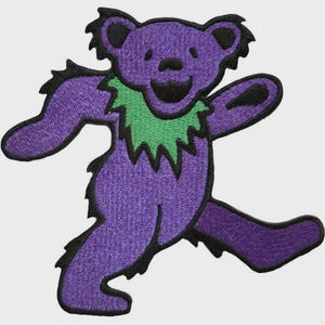 May include: A purple bear patch with a green collar and black outlines. The bear is in a dancing pose with its arms and legs outstretched. The patch is embroidered with a textured appearance.