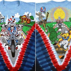 May include: A light blue tie-dye t-shirt featuring a colorful graphic design. The design includes a skeleton riding a motorcycle, a train, a house, and dancing bears. The shirt has a red, white, and blue V-shaped pattern.