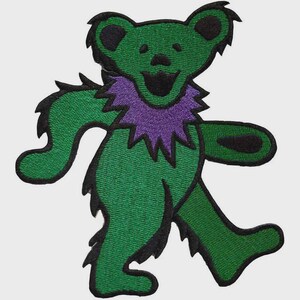 May include: Embroidered patch featuring a green bear with a purple collar and black accents. The bear is depicted in a walking pose with a smiling expression. The patch is likely for clothing or accessories.