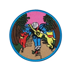 May include: A round, blue-bordered patch with an embroidered image of three cartoon bears walking on a path through a forest. The bears are red, yellow, and blue, and the bear in the middle is carrying a guitar. The background is a pink sky with green trees.