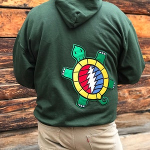 May include: Dark green hoodie with a large, colorful turtle graphic on the back. The turtle design features a yellow, red, and blue lightning bolt symbol within a green and yellow outline. The hoodie has a hood and is made of a soft material.