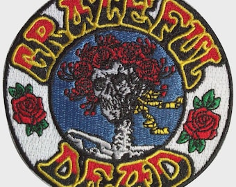 May include: A circular embroidered patch featuring a central skull figure adorned with red roses on a blue background. The text GRATEFUL DEAD is stitched in yellow and red lettering along the top and bottom edges, flanked by two red roses.
