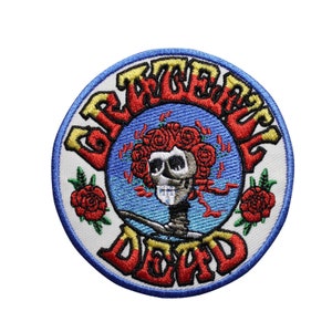 May include: Embroidered patch featuring the words "Grateful Dead" in red and yellow, encircling a skull with a crown of red roses. The patch has a blue border and two red roses on either side of the skull.