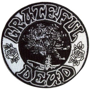 May include: A round, black and white embroidered patch featuring the words "Grateful Dead" in a stylized font. The center design includes a skull with roses and a skeleton. Two roses are on the outer edge.