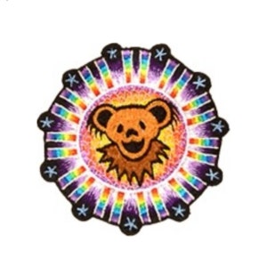May include: A circular patch featuring a smiling brown bear in the center, surrounded by a rainbow-colored sunburst design. The bear has a happy expression. The outer edge is a dark purple with white star-shaped accents. The patch is likely for clothing or accessories.
