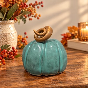 May include: A teal ceramic pumpkin ornament with a glossy, slightly distressed finish and a prominent, coiled metallic gold stem. The pumpkin features vertical ridges and a rounded shape, serving as a decorative object for seasonal displays.