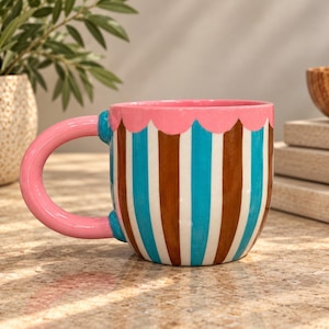 Könnte beinhalten: Ein Keramikbecher mit vertikalem Streifenmuster in Braun, Weiß und Hellblau. Der Becher hat einen gewellten rosa Rand und einen großen, abgerundeten rosa Henkel an der Seite. Die Oberfläche des Bechers hat ein glänzendes Finish.