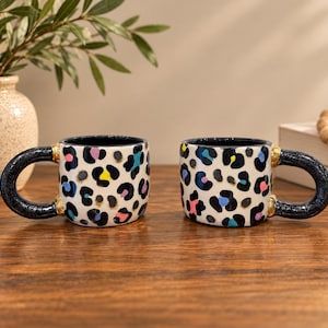 May include: Two ceramic mugs featuring a white base with a black leopard print pattern accented by small spots of blue, pink, and yellow. Each mug has a thick, dark blue handle with gold-colored rings where it attaches to the main body of the mug.