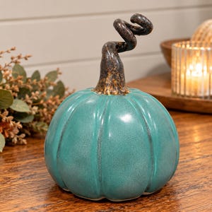 May include: A ceramic pumpkin figurine with a glossy teal finish and a dark, textured, spiraled stem. The pumpkin features vertical ridges and a smooth, rounded body, showcasing a distinct contrast between the vibrant teal base and the dark stem.