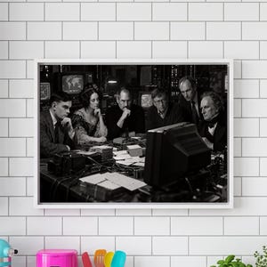 May include: A black and white framed wall art print depicting a group of historical and modern figures gathered around a desk with vintage computer equipment, including a monitor, cables, and papers, set in a dimly lit, cluttered workspace.