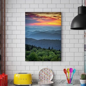 May include: A rectangular wall art print featuring a vibrant sunset over layered blue mountain ridges, with a foreground of dense green forest. The sky transitions from deep orange and yellow hues to soft purple tones above the distant peaks.