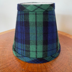 May include: A small lampshade featuring a traditional plaid design in green, blue, and black. The lampshade has a cylindrical form with a gentle flare and a rolled edge. It is placed on a wooden surface.