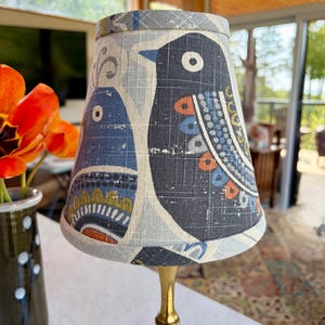 May include: A small lamp with a brass base and a fabric lampshade. The lampshade features a pattern of stylized blue birds on a gray background with accents of orange, white, and blue. The lamp is on a white surface.