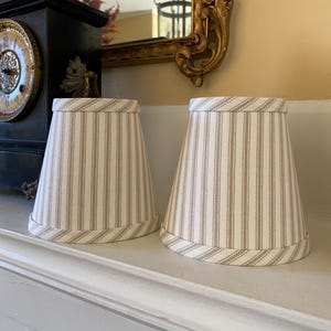 Puede incluir: Dos pantallas de lámpara beige con rayas marrones. Las pantallas de lámpara están hechas de tela y tienen un diseño simple y clásico.