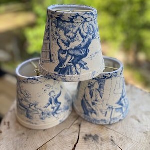 May include: Three white lampshades with a blue and white toile pattern featuring a scene of people in a garden. The lampshades are made of fabric and have a scalloped edge.