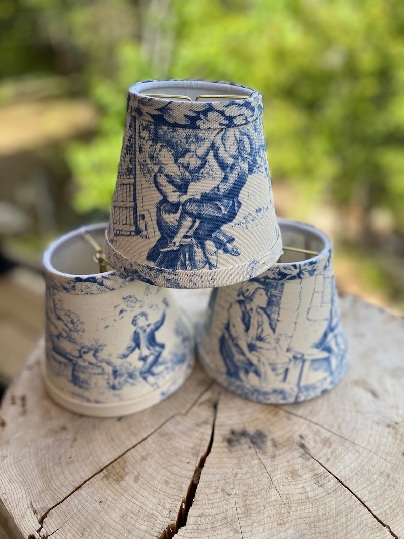 May include: Three white lampshades with a blue and white toile pattern featuring a scene of people in a garden. The lampshades are made of fabric and have a scalloped edge.