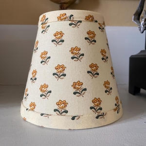 Puede incluir: Pantalla de lámpara de color crema con un patrón repetido de diseños florales naranjas y amarillos. La pantalla tiene una forma ligeramente acampanada y está rematada con un borde verde oscuro. Un accesorio de iluminación decorativo.