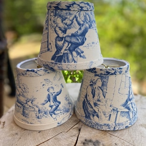 May include: Three blue and white patterned lampshades with a toile design. The lampshades are made of fabric and have a scalloped edge.