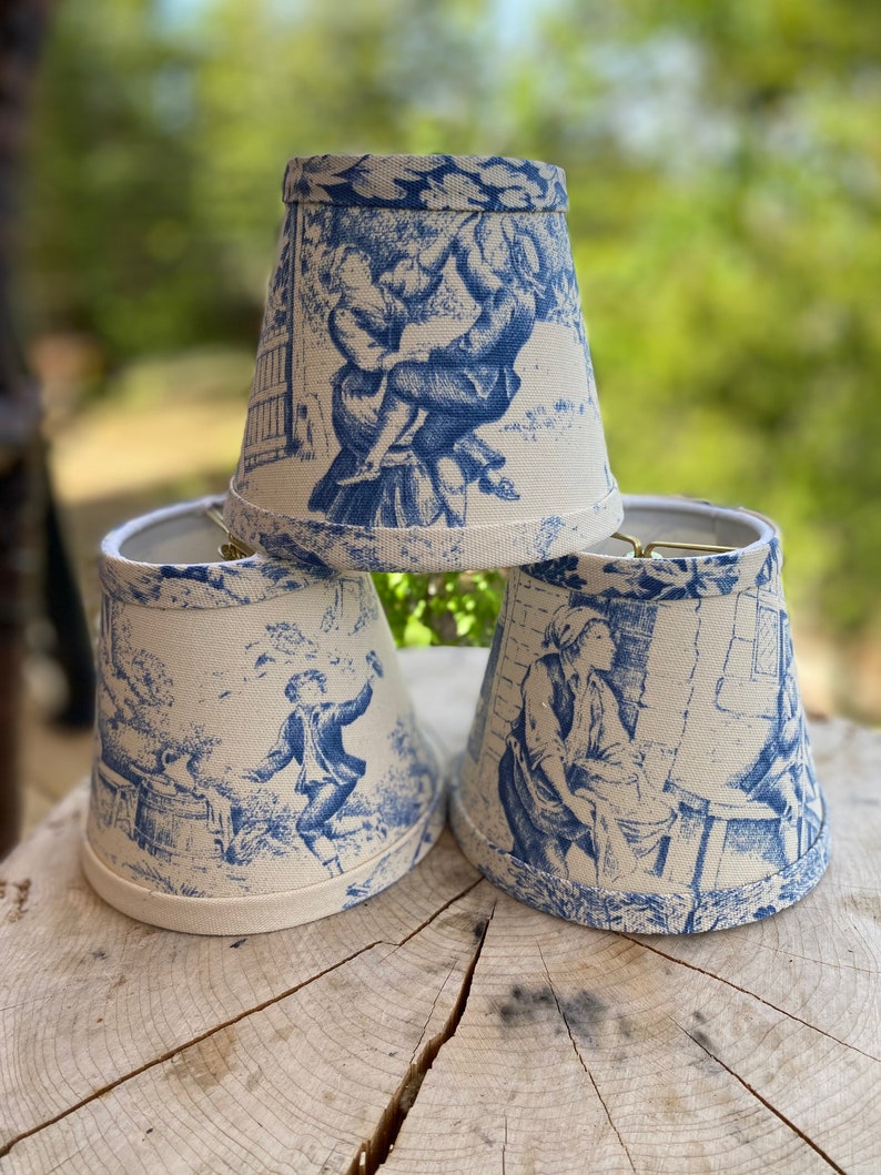 May include: Three blue and white patterned lampshades with a toile design. The lampshades are made of fabric and have a scalloped edge.