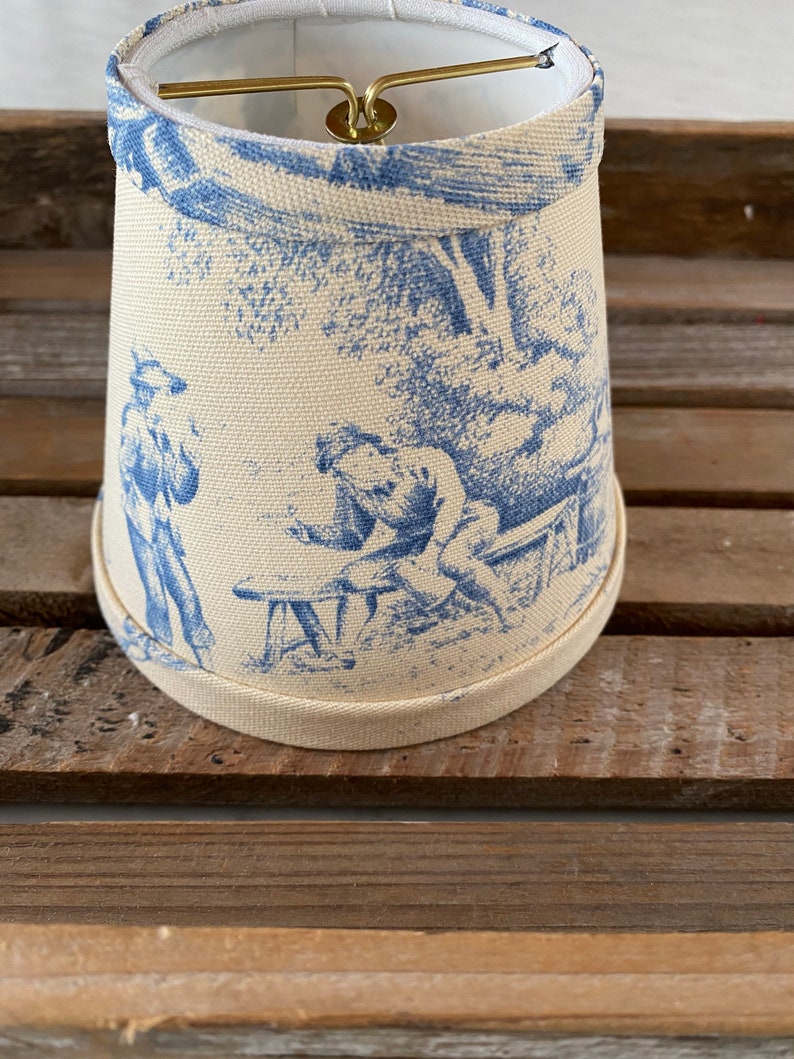 May include: A white lampshade with a blue and white toile pattern featuring a scene of two figures in a garden setting.