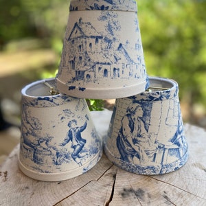 May include: Three white lampshades with a blue and white toile pattern. The lampshades are cone-shaped and have a scalloped edge.