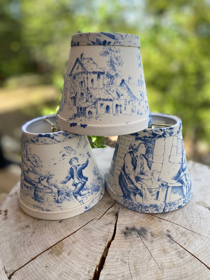 May include: Three white lampshades with a blue and white toile pattern. The lampshades are cone-shaped and have a scalloped edge.
