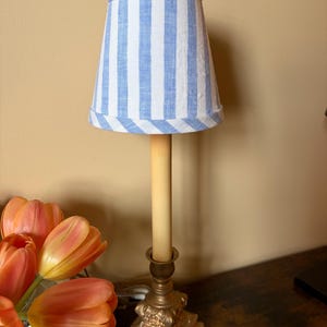 May include: A small table lamp with a blue and white striped lampshade. The lamp has a gold-colored base and a tall, cream-colored candle. A bouquet of orange tulips sits nearby on a dark wooden surface.