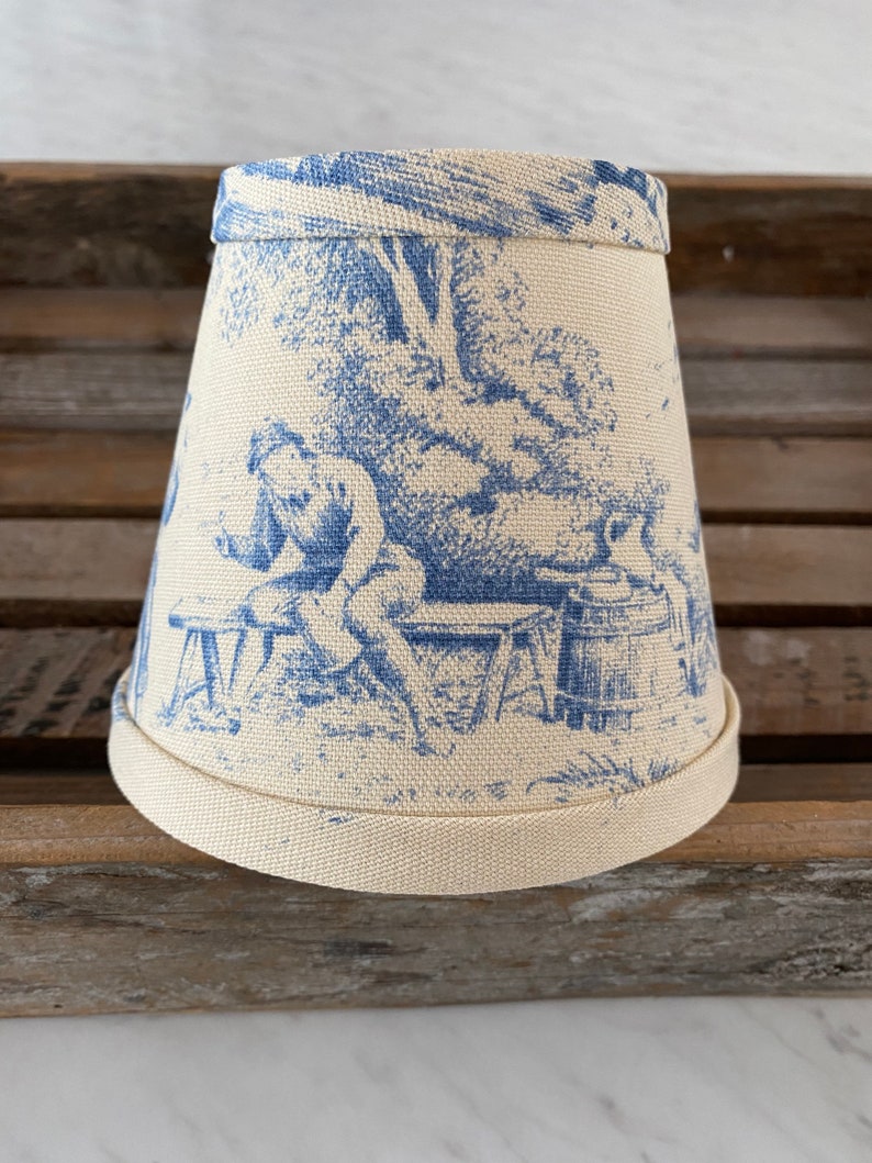 May include: A cream-colored lampshade with a blue toile pattern featuring a person sitting on a bench. The pattern includes trees and a small building.