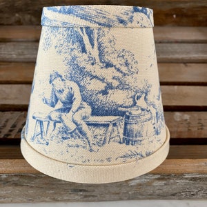 May include: A white lampshade with a blue toile pattern featuring a scene of a person sitting on a bench in a garden. The lampshade has a scalloped edge.