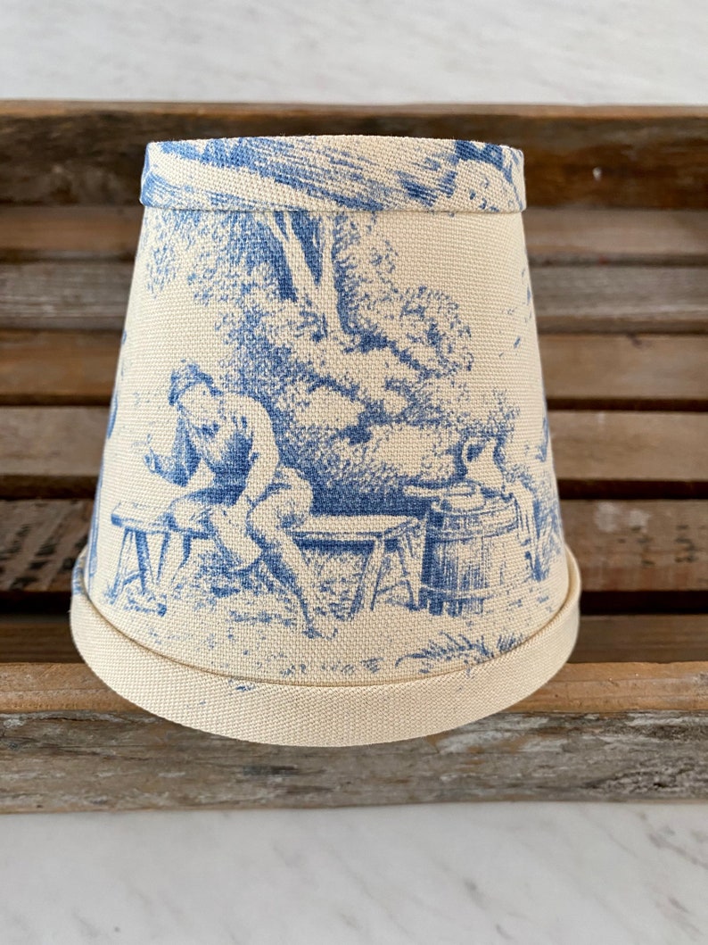 May include: A white lampshade with a blue toile pattern featuring a scene of a person sitting on a bench in a garden. The lampshade has a scalloped edge.