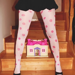 May include: White tights featuring a repeating pattern of pink polka dots, paired with a black skirt and black open-toe shoes. A small plastic toy house with a pink roof and tan walls is positioned on the wooden step between the legs.