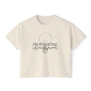 May include: A cream-colored short-sleeve t-shirt featuring a black line-art illustration of a lightbulb centered on the chest. The text "PROFESSIONAL idea-haver" is printed in a combination of serif and script fonts over the lightbulb graphic.