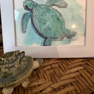 Könnte beinhalten: Ein gerahmtes Aquarellbild einer Meeresschildkröte in Grün- und Türkistönen mit dem Text Psalm 57:24 und einer Signatur. Neben dem Rahmen steht eine kleine, grüne Keramikschildkröte mit glänzender Oberfläche und detaillierter Panzerzeichnung.