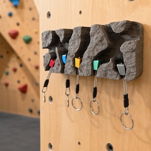 Puede incluir: Un llavero de pared con textura de roca gris que presenta cinco pequeñas presas de escalada en rojo, azul, amarillo, verde y plata. Cada presa está unida a un cable metálico con un anillo plateado para llaves colgando debajo.
