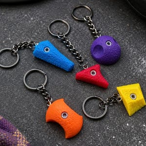 Puede incluir: Cinco llaveros con formas de presas de escalada en miniatura de colores azul, morado, rojo, naranja y amarillo. Cada pieza tiene una superficie texturizada, un orificio central con borde metálico y una cadena unida a una anilla metálica.