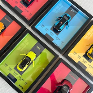 May include: A collection of framed shadow box displays, each featuring a miniature die-cast sports car mounted against a vibrant colored background. The displays include technical specifications like power, torque, top speed, and acceleration for models such as the Pagani, Lamborghini Huracán STO, BMW M4, and Bugatti Chiron.