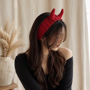 Puede incluir: Una diadema de punto de color rojo brillante que presenta dos cuernos de diablo puntiagudos y erguidos. La diadema tiene una textura acanalada y se lleva sobre la parte superior de la cabeza, mostrando una construcción de hilo hecha a mano.