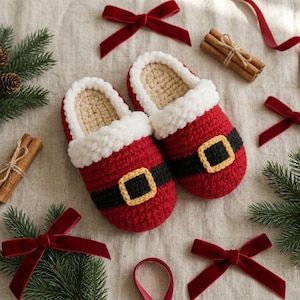 May include: A pair of red and white crochet slippers designed to resemble a Santa Claus suit, featuring a black belt with a yellow buckle, a white fluffy cuff, and a beige interior lining, arranged with pine branches and red velvet ribbons.