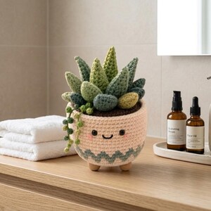 Peut inclure: Une plante succulente crochetée dans un pot rose clair avec un visage souriant, des joues roses et un motif en zigzag vert à la base. La plante présente des feuilles en laine de différentes nuances de vert et une petite tige tombante.