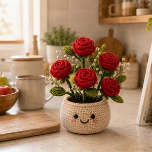 Könnte beinhalten: Eine gehäkelte Topfpflanze mit fünf leuchtend roten Rosen, grünen Blättern und kleinen weißen Akzentblüten. Der runde, cremefarbene Häkeltopf hat ein aufgesticktes lächelndes Gesicht mit schwarzen Augen, einem Mund und rosa Wangen.