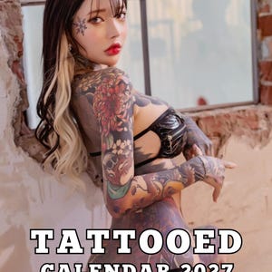 May include: A 2027 calendar featuring a person with extensive colourful tattoos on their arms and body, wearing a black top. The text TATTOOED CALENDAR 2027 is printed in white with a black outline at the bottom, followed by smaller text in four languages.