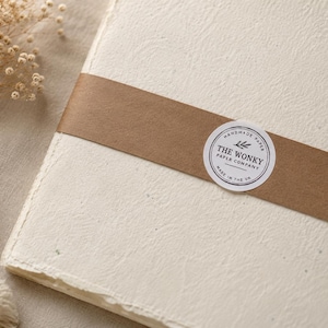 May include: A stack of off-white, textured handmade paper sheets secured with a brown paper band. A circular white sticker on the band features the black text THE WONKY PAPER COMPANY, a small leaf icon, and the words HANDMADE PAPER MADE IN THE UK.