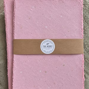 May include: A stack of rectangular sheets of light pink handmade paper with deckled edges. A brown paper band wraps around the centre, featuring a circular white label with the text "HANDMADE PAPER THE WONKY PAPER COMPANY MADE IN THE UK".