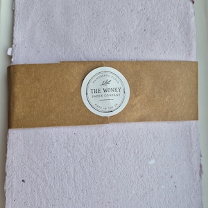 May include: A stack of rectangular, light lavender handmade paper with a textured, fibrous surface. A brown paper band wraps around the center, featuring a circular white sticker with the text "HANDMADE PAPER THE WONKY PAPER COMPANY MADE IN THE UK".