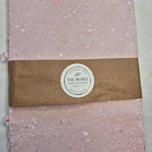 May include: A stack of rectangular handmade paper with a light pink colour and speckled texture. A brown paper band wraps around the centre, featuring a circular white label with the text "HANDMADE PAPER THE WONKY PAPER COMPANY MADE IN THE UK".