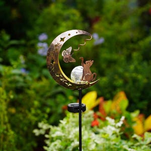 May include: A solar garden stake featuring a bronze-colored crescent moon cutout with star shapes, a metal butterfly, and a silhouette of a cat reaching toward a clear, crackled glass orb light mounted on a thin black metal pole.